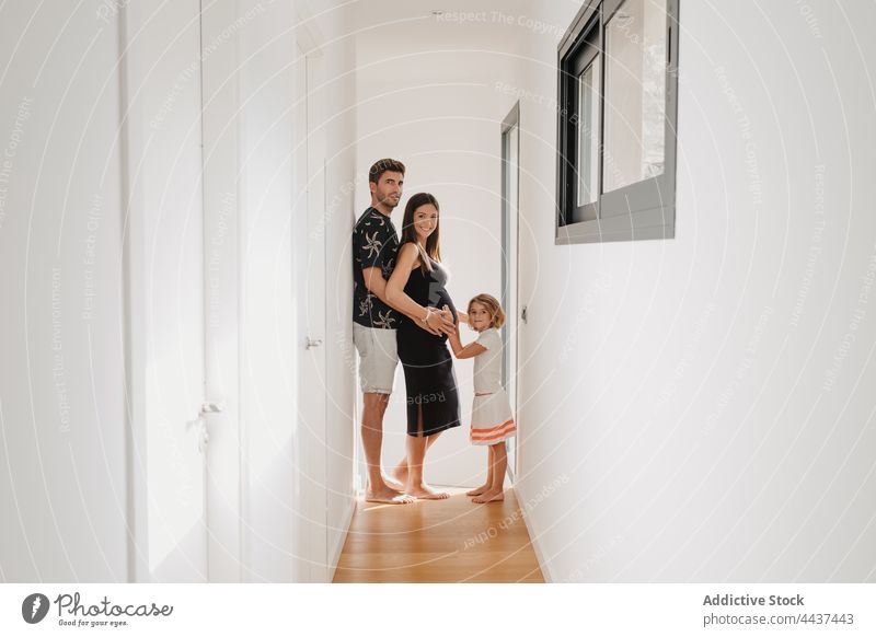 Girl embracing tummy of pregnant mom against father in corridor family pregnancy embrace motherhood expect smile fondness house portrait barefoot stand passage