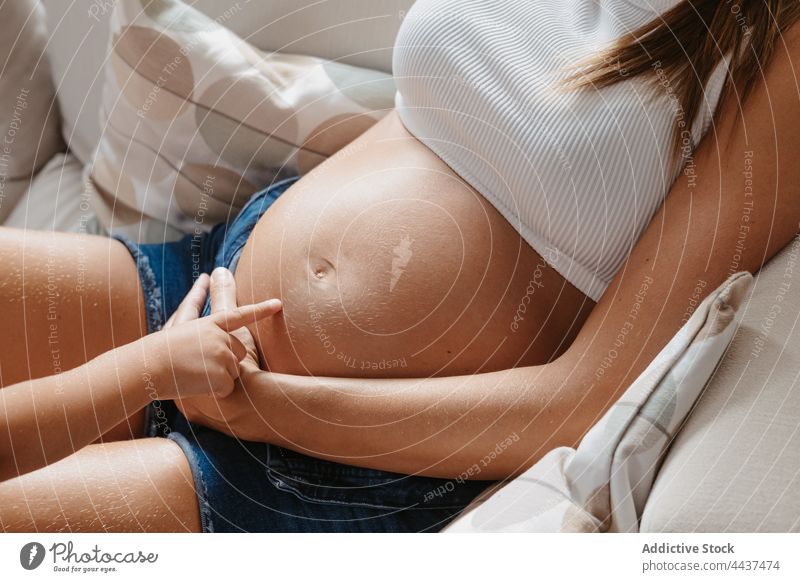 Anonymous pregnant mother and daughter on sofa interact pregnancy stroke spend time childhood caress motherhood fondness belly home woman kid girl barefoot