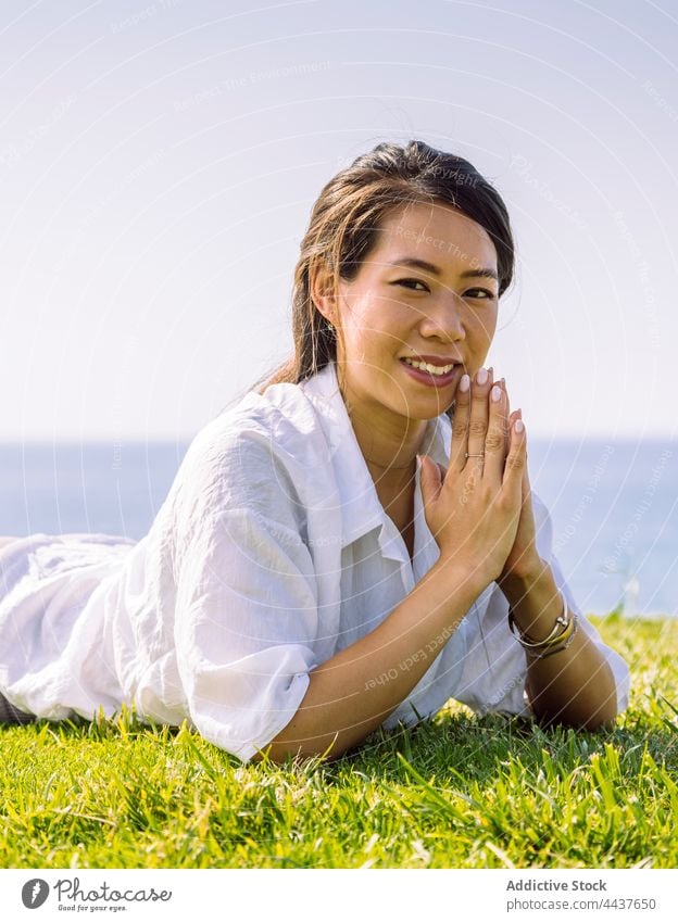 Charming Asian woman with clasped hands resting on grass shore smile hands clasped sincere charming friendly pleasant seashore portrait kind idyllic gaze candid