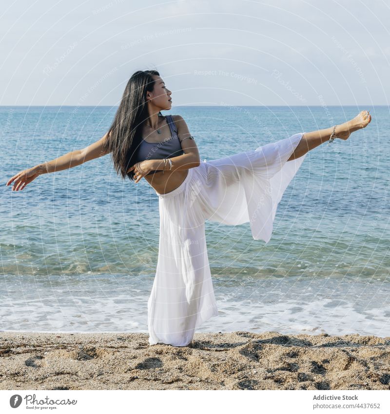 Asian woman with raised leg practicing yoga on beach leg raised balance practice healthy lifestyle stretch wellness vitality seashore asian ethnic trousers