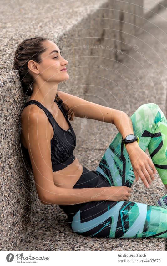 Delighted sportswoman sitting near wall in city runner break athlete cheerful smile training pause fit female summer sportswear workout lean vitality happy