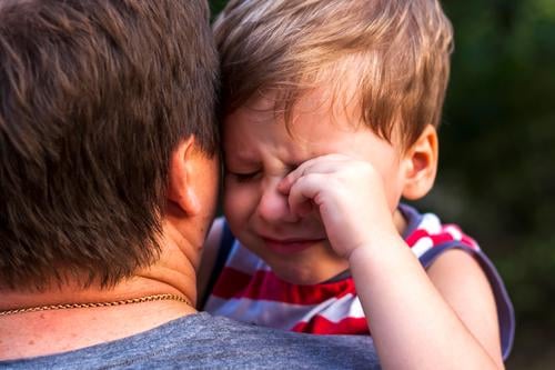 A little boy is crying while being in his father's arms, snuggling up to him to feel protected. expression caring relationship child son white parent sitting