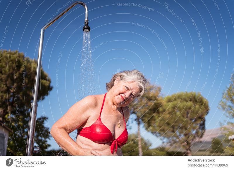 Smiling woman standing under shower on poolside splash water relax enjoy wash clean female transparent drop cheerful pleasure senior old vacation sunlight blue