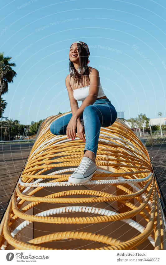 Smiling ethnic woman squatting on geometric construction in town trendy cool individuality cheerful enjoy architecture geometry street millennial headset