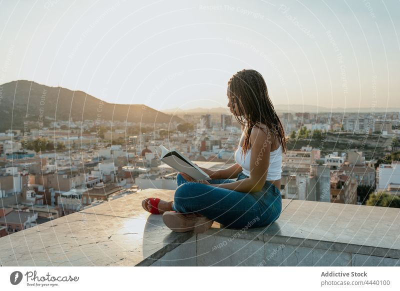 Cool black woman reading book on rooftop in city textbook spare time literature trendy legs crossed attentive sandal creative design footwear focus millennial