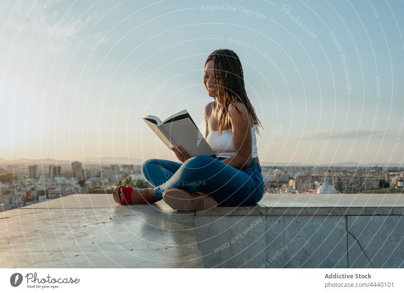 Cool black woman reading book on rooftop in city textbook spare time literature trendy legs crossed attentive sandal creative design footwear focus millennial