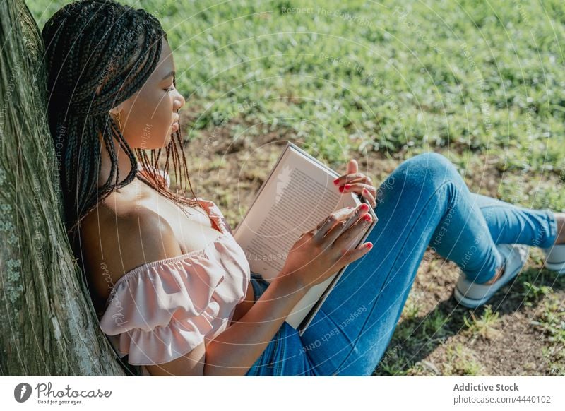 Contemplative ethnic woman with book resting in park contemplate wistful reflective trendy millennial lawn gumshoe crop top textbook literature trunk meadow