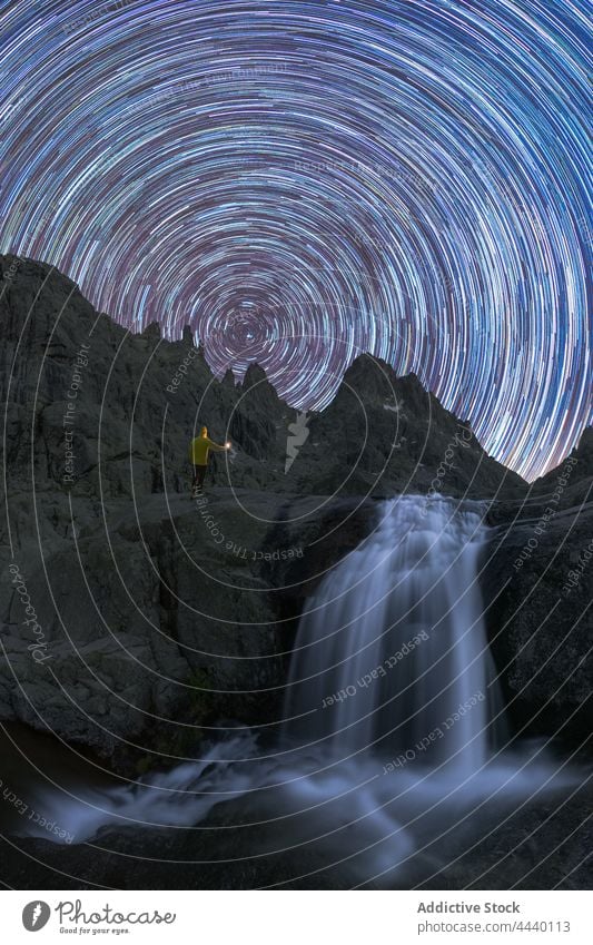 Tourist contemplating waterfall on mountain under star trail at night traveler cascade nature highland motion speed energy dynamic foamy pond fast power starry