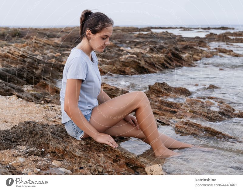 Teenage girl sitting on cliff by the sea teenager blue Caucasian rock sunset adolescent stone wearing childhood female copy space jeans lifestyle alone casual