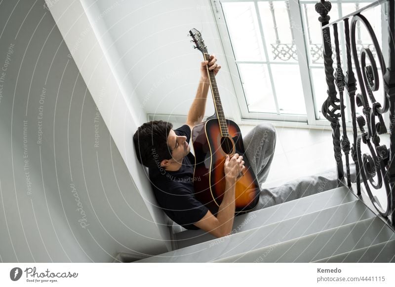 Relaxed young man playing guitar on staircase near window at home. music instrument acoustic guitarist hobby male skill practice alone compose rest relax melody