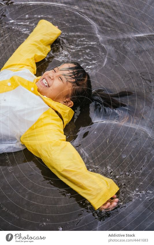 Happy ethnic girl in raincoat having fun in puddle carefree laugh legs apart arms apart childhood pastime rainy spare time reflection pavement gumboots jeans