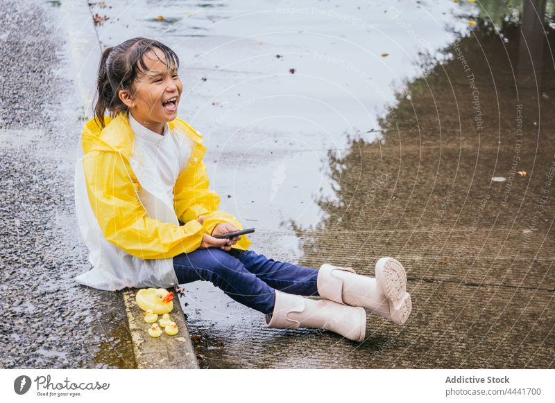 Happy Asian girl with smartphone on pavement in rainy weather laugh carefree childhood toy puddle rest gumboots having fun smile pastime duck weekend spare time