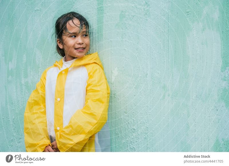 Smiling Asian girl standing against rough wall rain cheerful childhood enjoy raincoat sincere charming asian smile ethnic content ponder candid charismatic