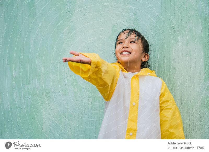 Smiling Asian girl catching rain drops against rough wall palm up cheerful childhood enjoy raincoat sincere charming asian smile ethnic content ponder candid
