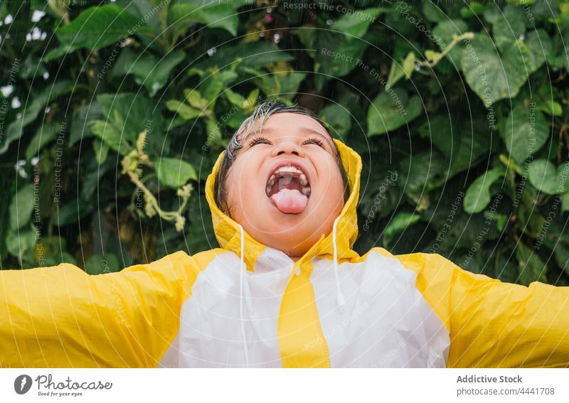 Happy Asian girl drinking rain against bush in park tongue out mouth opened having fun childhood cheerful carefree enjoy shrub friendly sincere kind raincoat