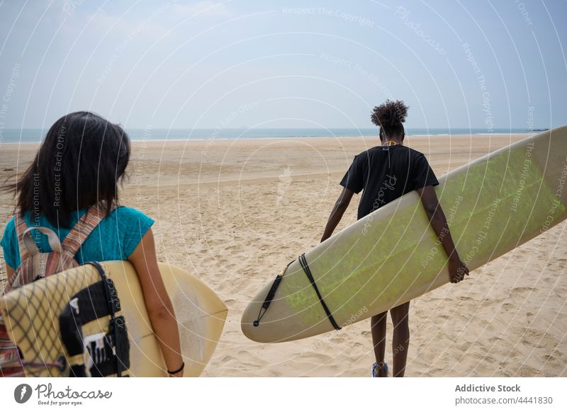 Diverse sportswomen with surfboards talking while walking on ocean coast friendship surfing spend time shore blue sky smile surfer speak longboard black content