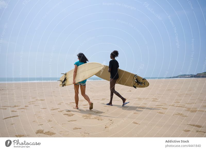 Diverse sportswomen with surfboards talking while walking on ocean coast friendship surfing spend time shore blue sky smile surfer speak longboard black content