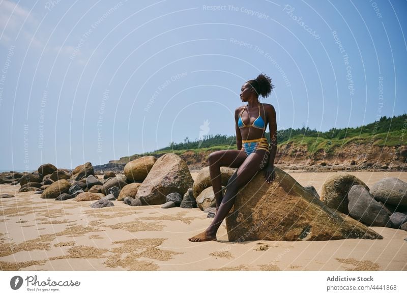 Smiling black woman in swimwear resting on boulder on coast body smile feminine gentle afro natural seacoast cheerful beauty seashore blue sky content charming