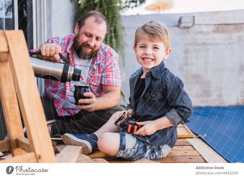 Father serving mate against smiling son with wooden blocks father pour smile thermos woodwork lumber cheerful attentive diy break man dad happy herbal tea