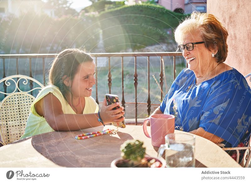 Positive girl showing smartphone to cheerful grandmother on terrace in sunlight granddaughter happy smile demonstrate woman child share table relationship