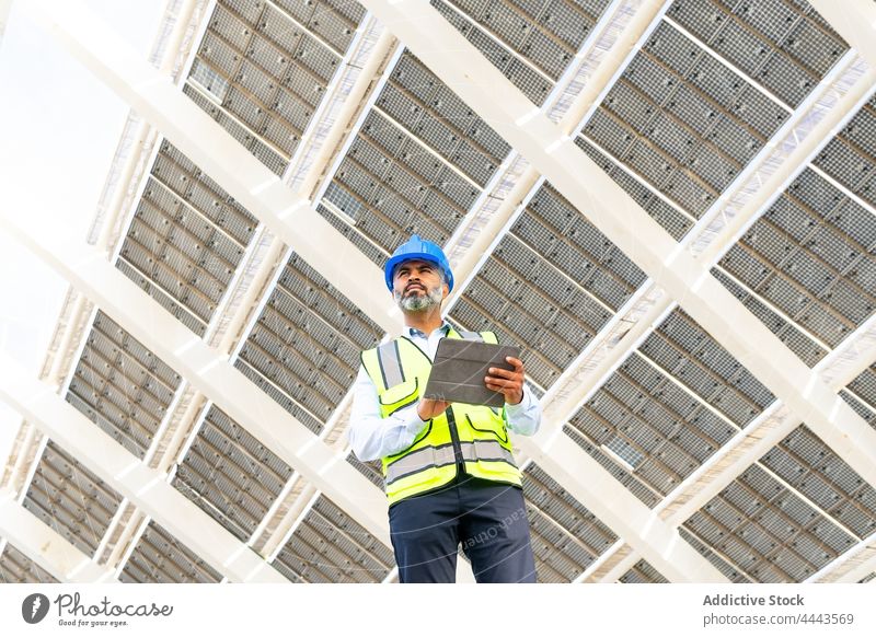 Ethnic contractor with tablet standing near solar power station man work professional using gadget device ecology foreman uniform modern photovoltaic