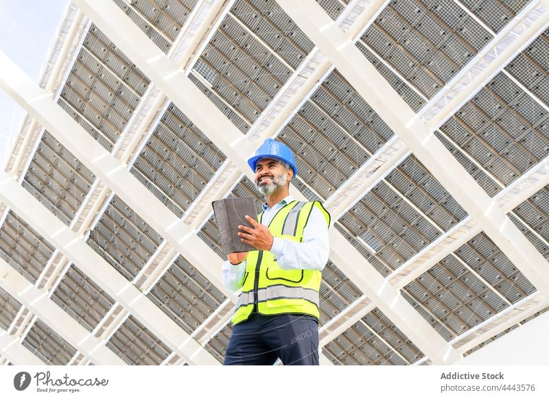 Smiling ethnic contractor with tablet standing near solar power station man work professional smile happy using gadget device ecology foreman uniform modern