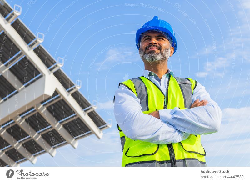 Anonymous engineer with hardhat against solar power station in sunlight man safety profession vest blue sky foreman modern arms crossed sustainable technician