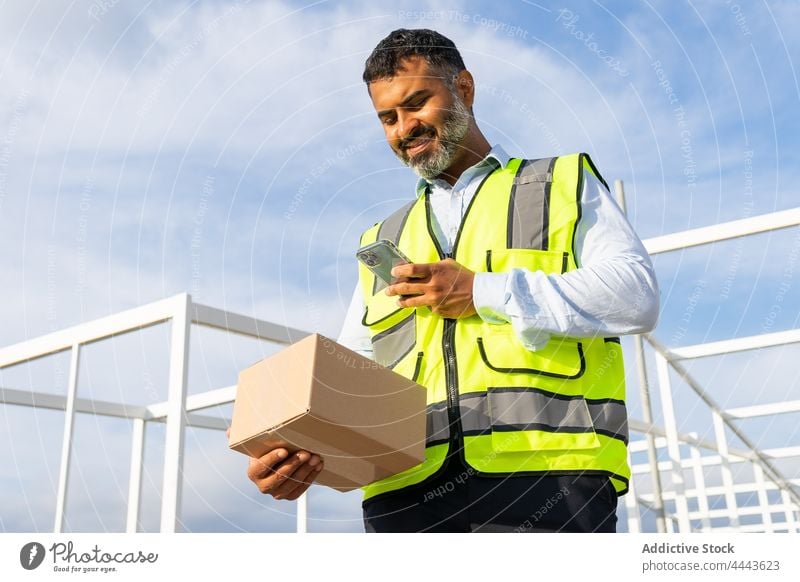 Smiling ethnic workman with box browsing smartphone worker smile happy using carry parcel glad delivery uniform delight watch male occupation job device gadget
