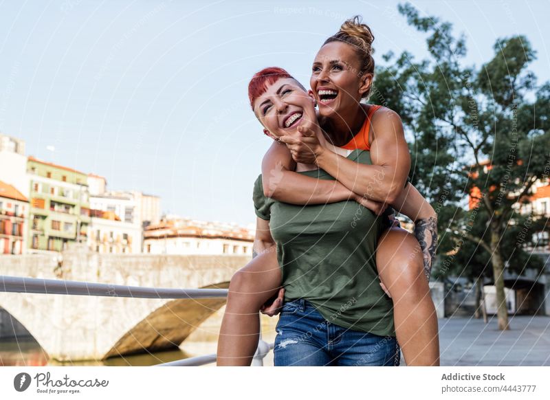 Happy woman carrying homosexual girlfriend piggyback on embankment having fun laugh relationship carefree cool same sex town women embrace love lgbt smile