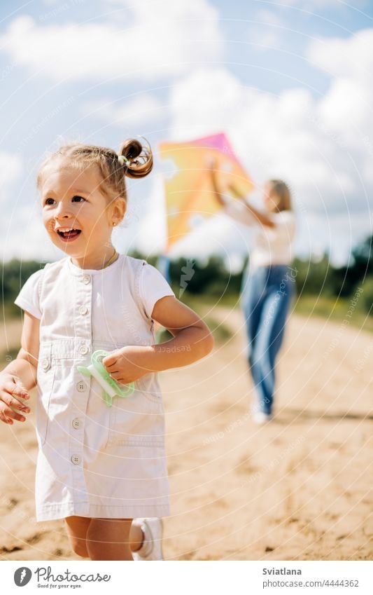 A little girl in a white dress is running along the road with a string of a kite in her hand, a girl is flying a kite with her mother mom baby launching