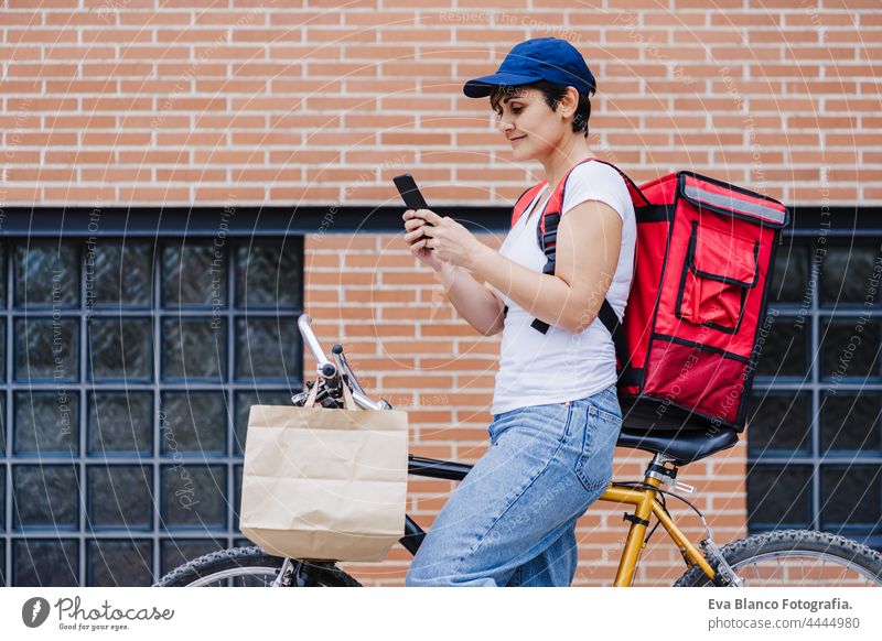 rider woman wearing red backpack delivering food on a bike, checking order with smart phone while standing on street in city. Delivery service concept. Sustainable transport