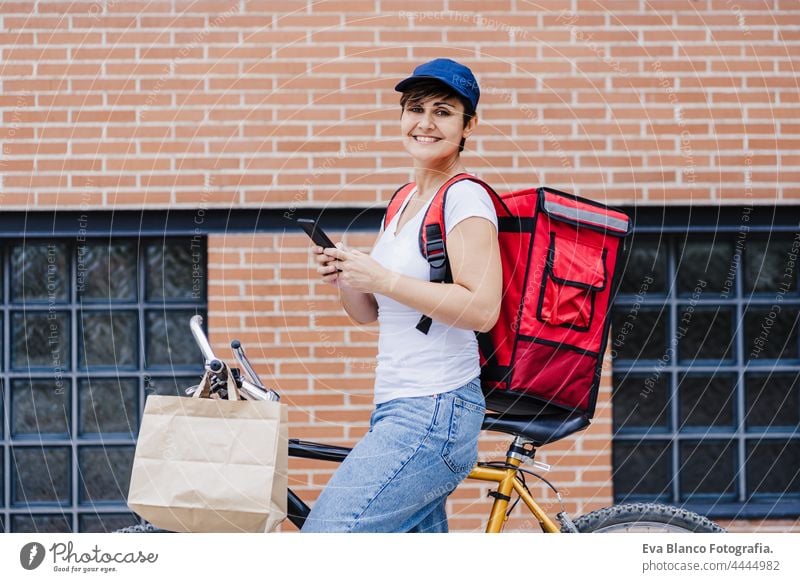 smiling rider woman wearing red backpack delivering food on a bike, checking order with smart phone while standing on street in city. Delivery service concept. Sustainable transport