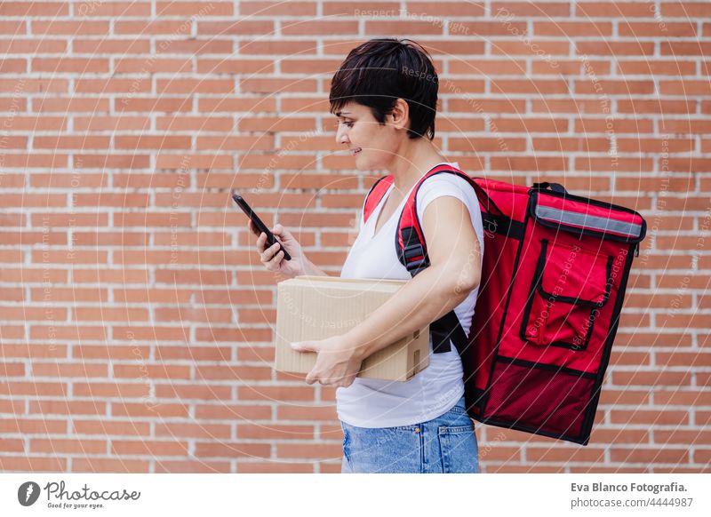 happy young rider woman wearing red backpack holding paperboard box of food in city. Delivery service concept. Woman checking order in mobile phone deliver