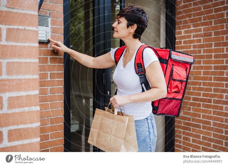 young rider woman wearing red backpack holding paperboard box of food in city. Delivery service concept. Woman using an intercom system deliver bicycle home