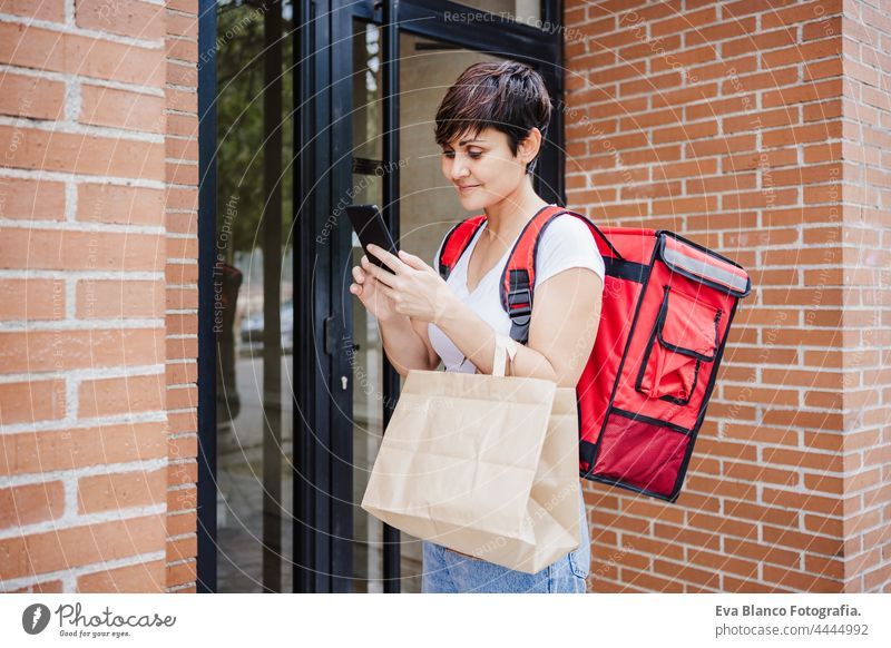 young rider woman wearing red backpack holding paperboard box of food in city. Delivery service concept. Woman checking order in mobile phone deliver bicycle