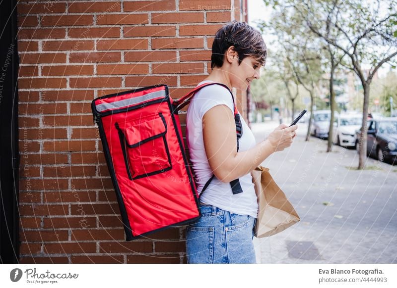 side view of young rider woman wearing red backpack holding paperboard bag of food in city. Delivery service concept. Woman checking order in mobile phone