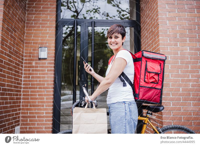 smiling young rider woman wearing red backpack holding paperboard bag of food in city. Delivery service concept. Woman checking order in mobile phone deliver