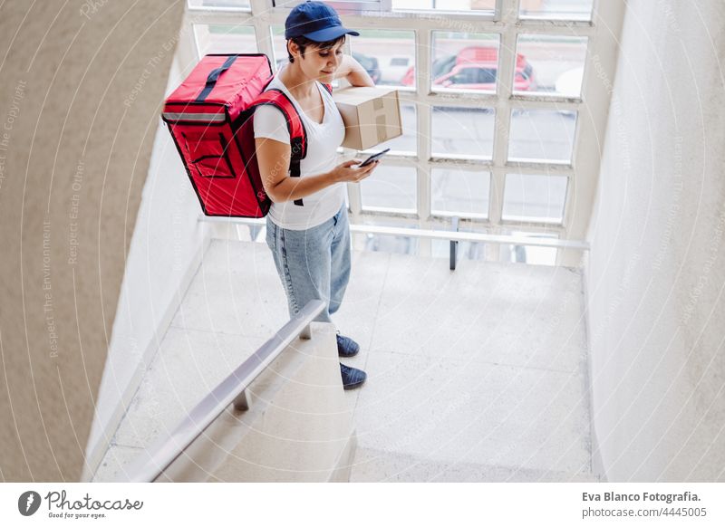 young rider woman wearing red backpack holding paperboard box in neighborhood stairs. Delivery service concept. Sustainable transport deliver food bicycle home