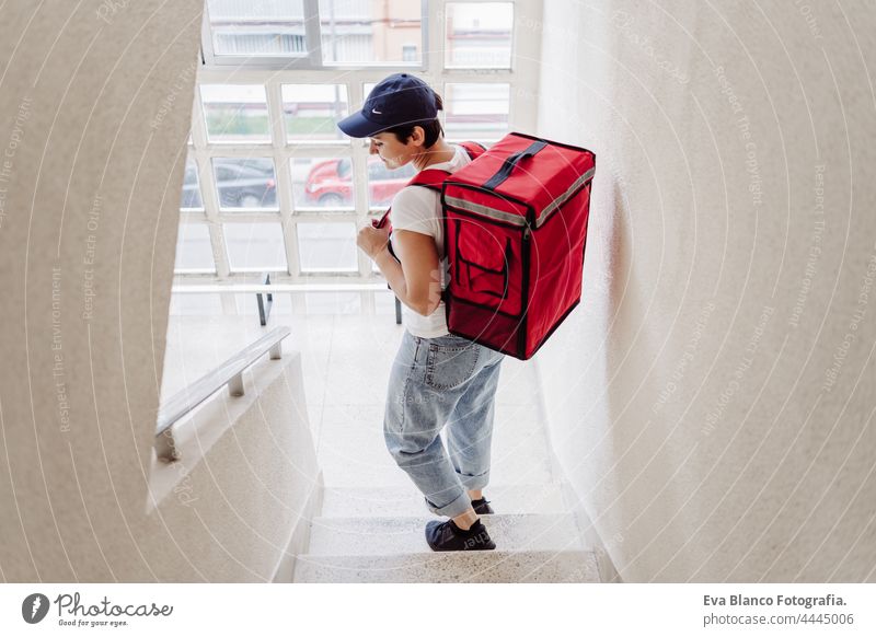 back view of young rider woman wearing red backpack holding paperboard box in neighborhood stairs. Delivery service concept. Sustainable transport deliver food