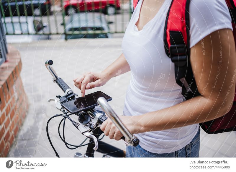 close up of Young rider woman wearing red backpack delivering food on a bike, checking order with smart phone while standing on street gate in city. Delivery service concept. Sustainable transport