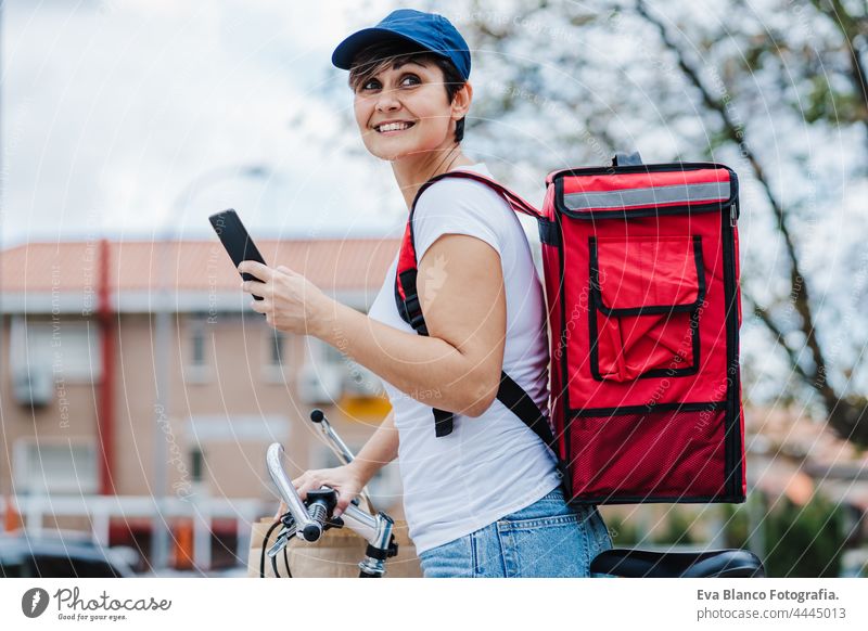 happy rider woman wearing red backpack delivering food on a bike, checking order with smart phone while standing on street in city. Delivery service concept. Sustainable transport
