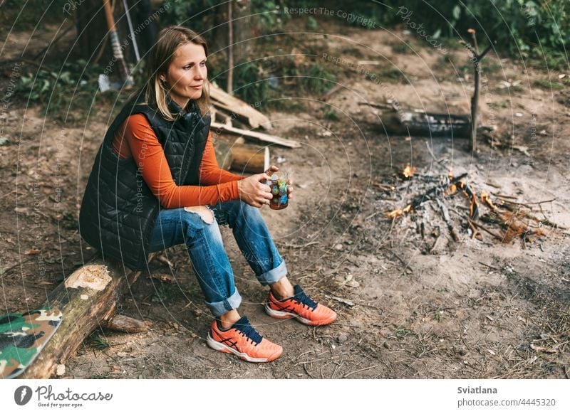 A young woman is warming herself by the fire with a cup of warming tea in the forest drink sitting nature tourist adventure vacation campfire bonfire travel
