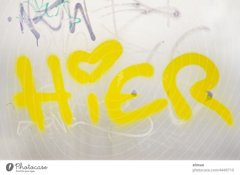 HERE is written in bright yellow letters and with a heart as an i-dot on the grey metal wall / graffito here Yellow location Meeting point Graffito Graffiti