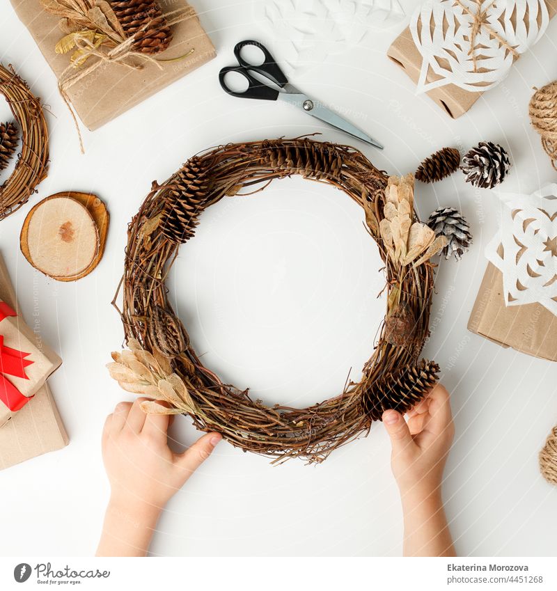 Child's hands making Christmas wreath. Flat lay, top view, Kid hands wrapped gift box in craft paper, natural decoration details, zero waste packaging on white table. Eco-friendly concept
