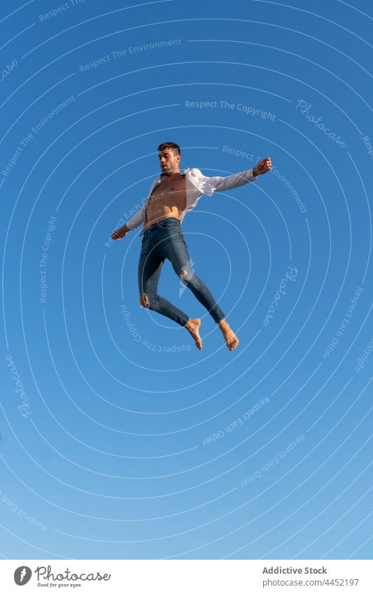 Active dancer jumping against blue sky trick active energy dynamic fearless man extreme leap barefoot trendy wear jeans white shirt perform brave activity