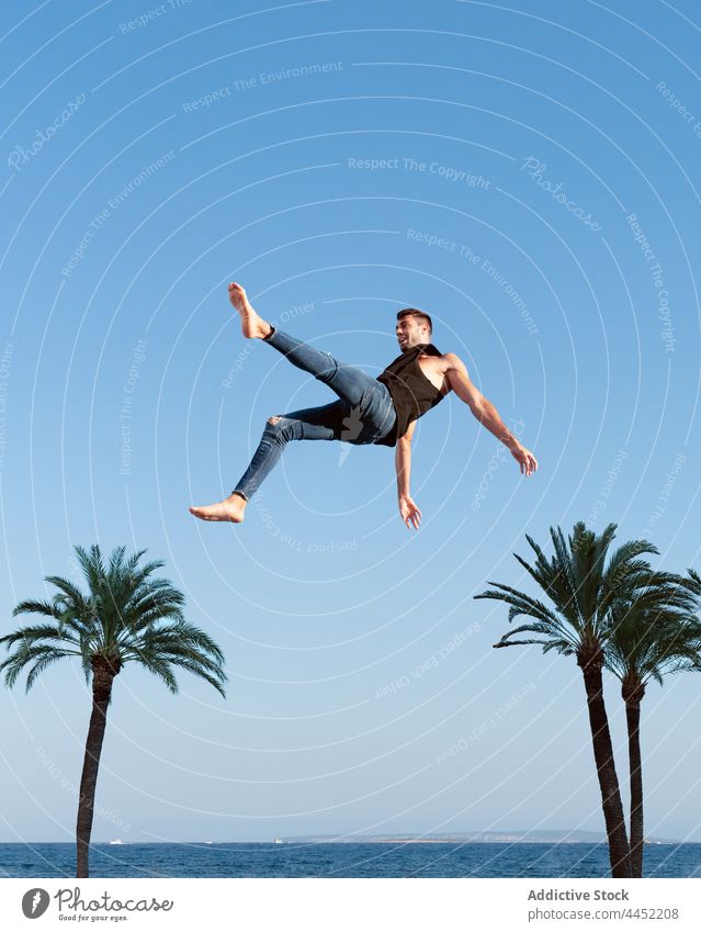Active dancer jumping against palm tree under blue sky trick active energy dynamic fearless man extreme leap barefoot trendy wear jeans white shirt perform
