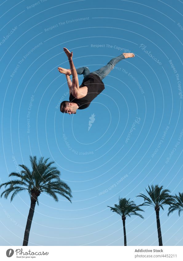 Energetic dancer performing trick in mid air above palm trees backbend jump extreme energy courage adrenalin blue sky man dynamic fearless nature touch leg