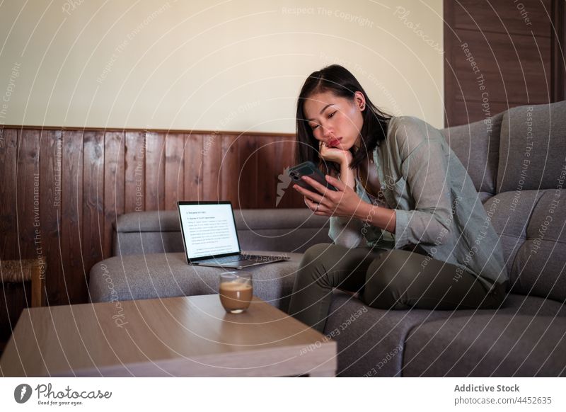 Asian woman with latte touching screen on smartphone touch screen surfing internet coffee hot drink home using gadget browsing aroma focus sofa house table