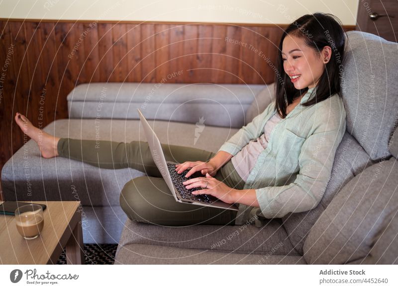Asian freelancer typing on laptop on couch against latte work internet project smile sofa woman using happy gadget interested device coffee smartphone hot drink