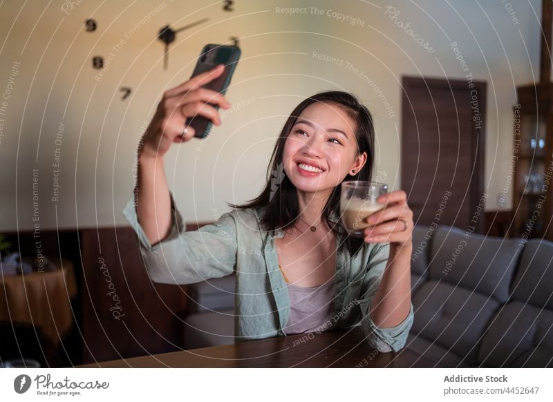Smiling Asian woman taking selfie with latte on smartphone smile laptop coffee sofa house using gadget spare time cheerful content self portrait device